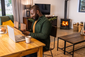 African American man sitting at dining table checking phone with silver laptop and mug on coaster
