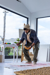 Indian man crouching over floor canvas in art studio, holding wooden mixing bowl and wearing beanie