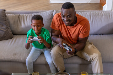 African American father and son sitting on gray sofa holding wireless gamepads near glass table © wavebreak3