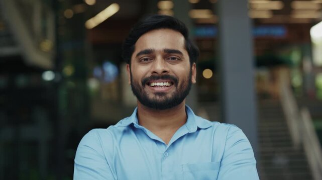 Portrait of positive Indian guy in blue shirt smiles near shopping mall. Young Hindu man in casual attire radiates positive emotions in urban old district. Corporate employee