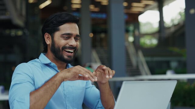 Portrait of smiling Indian entrepreneur talks on video call with sign language via laptop in cafe. Friendly man uses gestures for words at online meeting outdoor corporate office