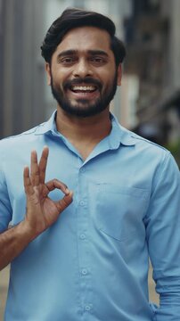 Portrait of joyful Indian guy shows Ok gesture as stands on street vertical shot. Happy bearded businessman expresses agreement with hand sign in old town. Body language symbol
