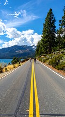Naklejka premium A serene road with double yellow lines stretching into the distance, flanked by trees and a body of water under a blue sky