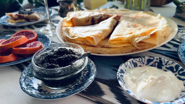 Luxury Maslenitsa breakfast: Russian crepes with black caviar, white wine and pink tulips. Elegant table setting with a black cat among houseplants on the background. Expensive lifestyle concept.