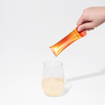 Hand pouring vitamin or collagen powder from orange sachet into glass of water