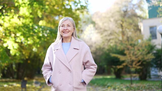 Smiling senior woman in a coat walking through an urban city park, enjoying a pleasant day outdoors surrounded by green trees, feeling relaxed and joyful while appreciating nature and fresh air.