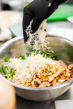 A chef in black gloves sprinkles freshly grated Parmesan cheese over a large Caesar salad featuring grilled chicken and crisp romaine lettuce in a stainless steel mixing bowl.