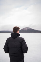 Fototapeta premium View of snowy hill across Macha Lake with blurred man in foreground. Winter landscape in Czech Republic. Man standing near frozen Macha Lake in Czech Republic looking at distant hill. Winter travel an