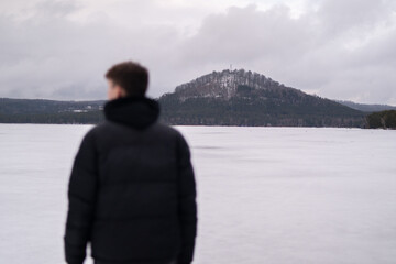Fototapeta premium View of snowy hill across Macha Lake with blurred man in foreground. Winter landscape in Czech Republic. Man standing near frozen Macha Lake in Czech Republic looking at distant hill. Winter travel an