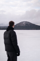 Fototapeta premium View of snowy hill across Macha Lake with blurred man in foreground. Winter landscape in Czech Republic. Man standing near frozen Macha Lake in Czech Republic looking at distant hill. Winter travel an