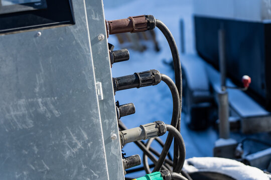 Multiple heavy duty cable connectors attached to metal control box, black cords looping in cold conditions with blurred machinery behind, industrial electrical hookup and field service theme