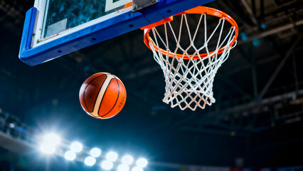 Fototapeta premium Basketball ball flying into hoop net on indoor court during game with bright stadium lights representing active sport and competitive leisure time.