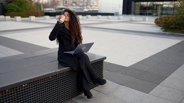 Young businesswoman with curly hair working on her laptop while sitting on a bench in a modern city square and drinking coffee