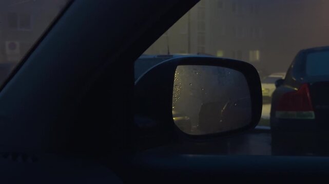 Rainy evening wait. Raincoated mirror shows blurred city scene. Evening rain reflection captures driver waiting patiently outside