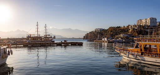 Naklejka premium Scenic view of Antalya old town harbor Kaleici at bright winter sunset. Coastal landscape with excursion boats, mediterranean sea, rocky cliffs and mountains in haze. Antalya, Turkey.