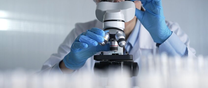 Woman scientist wearing lab coat, white protective gloves and glasses, is using microscope in modern laboratory setup, portrait view. Low key lighting photo. Science and Medicine