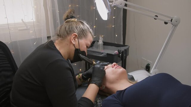 Beauty procedure: relaxed young girl gets permanent eyebrow tattoo with needle machine for natural look. Master holds brow with fingers during microblading in beauty salon, high detail slow motion.
