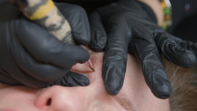 Close-up salon treatment: woman receives microblading permanent eyebrow tattoo with needle machine. Beautician corrects shape with fine strokes in beauty salon, natural result.
