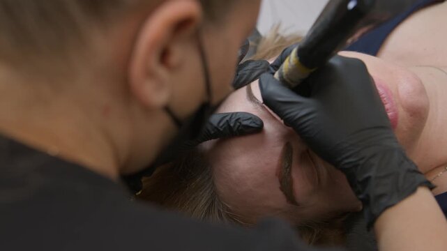 Detailed close-up: permanent eyebrow tattoo microblading session in beauty salon. Beautician with needle machine works on woman, creating realistic hair-like brows, professional beauty correction 