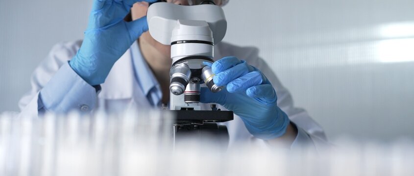 Woman scientist wearing lab coat, white protective gloves and glasses, is using microscope in modern laboratory setup, portrait view. Low key lighting photo. Science and Medicine