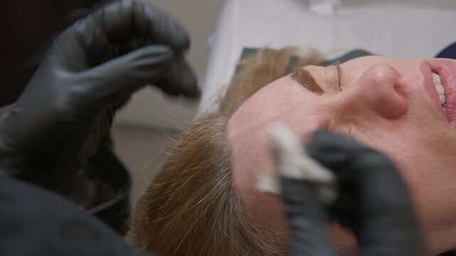 Close-up of microblading procedure: master uses special needle tattoo machine to tattoo permanent eyebrows on woman in beauty salon. Fine artistic strokes for beautiful natural shape, high detail 4K.