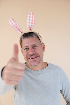 Man with grey hair and brown sweater with Easter bunny ears standing in front of brown background