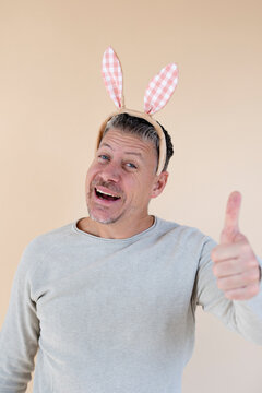 Man with grey hair and brown sweater with Easter bunny ears standing in front of brown background