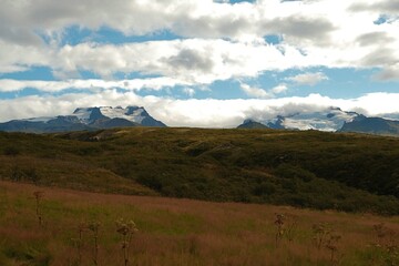 Obraz premium Wide Icelandic landscape with grassy hills and distant glaciers of Vatnajokull under dramatic clouds.