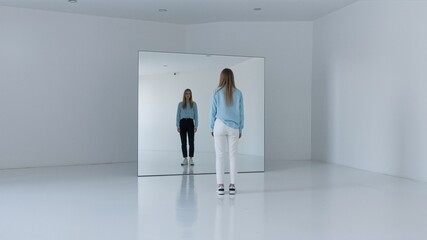Fototapeta premium Young woman in blue shirt stands facing her reflection in a mirror
