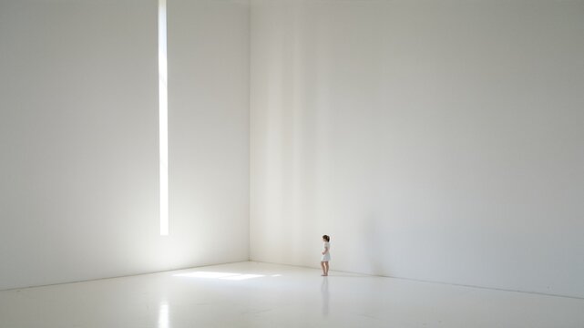 Young girl in white dress standing in a minimalist white room with light