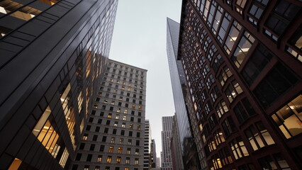 Naklejka premium low angle cityscape at dusk showing warm office lights, brick and glass facades, narrow canyon street, subtle reflections, calm mood ideal for architecture,