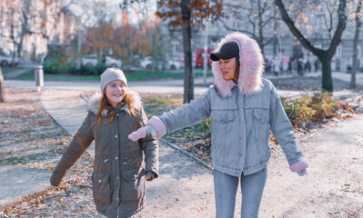 Obraz premium Happy mother and ten year old daughter smiling and playing together in sunny autumn urban park while holding hands with soft bokeh background and warm lens flare on bright day with copy space