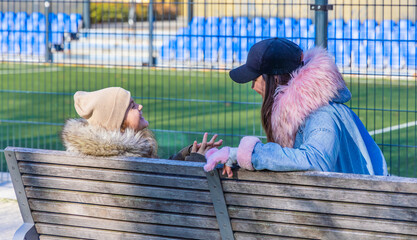A smiling mother and her teenage daughter sitting together on a wooden park bench near a green soccer pitch with blue stadium seating, posing while waiting for a game on a sunny autumn day © Igor