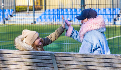 Happy mother and ten year old daughter giving a high five while sitting on a wooden bench near a soccer field with blue stadium stands in a sunny urban park with copy space and soft bokeh background © Igor