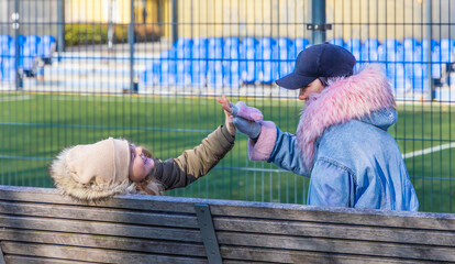 Happy mother and ten year old daughter giving a high five while sitting on a wooden bench near a soccer field with blue stadium stands in a sunny urban park with copy space and soft bokeh background © Igor