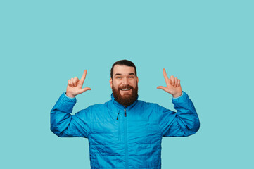 Fototapeta premium Playful Caucasian Man Pointing Upward With Smile, Bright Turquoise Background And Blue Jacket Conveying