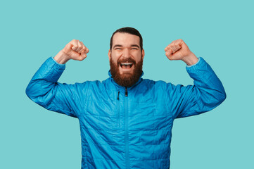Fototapeta premium Triumphant Caucasian Man With Raised Fists, Joyful Expression Against Turquoise Backdrop Wearing Blue Jacket,
