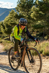 Woman cycling on a gravel bike during an outdoor adventure, traversing a rural mountain road with pine trees and hills under a partly cloudy sky in alicante, costa blanca - stock photo