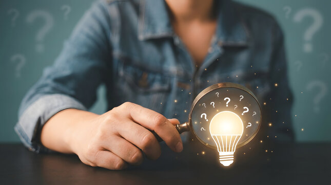 Close up of a person holding a magnifying glass over a glowing light bulb surrounded by question marks symbolizing the search for answers and innovative solutions