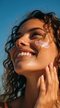 Close up of a happy young woman with curly hair and freckles wearing sunscreen cream on her face outdoors under a clear blue sky (Vertical Video)