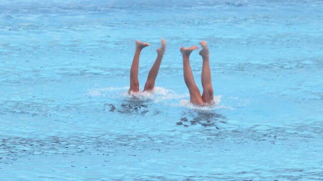 Two artistic swimmers performing synchronized leg movements during a duet routine in a swimming pool