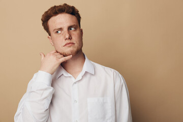 Fototapeta premium Young man with red hair and freckles in white shirt makes call me gesture with hand near face, looking sideways on beige background. Concept of communication, gesture, and casual style.