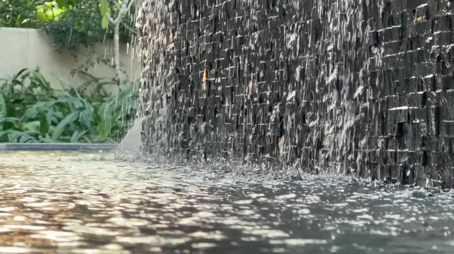 Water flows down a vertical fountain wall, forming a smooth water curtain over stone surface beside a small pool and garden greenery. Decorative fountain.
