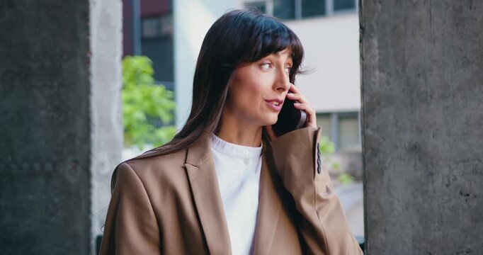 Smiling self employed woman making phone call on mobile phone and taking notes while working remotely while sitting on bench about modern office building, outdoors