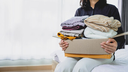 Woman holding box of folded clothes for donation while sitting on bed. Representing charity, clothing donation, recycling and helping people concept.