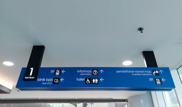 Blue hospital wayfinding sign in Bahasa Indonesia and English language with symbols, icons, and arrows directing to clinic, elevators, toilets, in-patient admission, and information desk in the lobby.