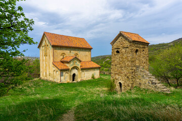 Fototapeta premium Stone church with tiled roof and stone tower in the courtyard of Maghlaani church complex. Green grass. Mountains and sky with clouds in the background