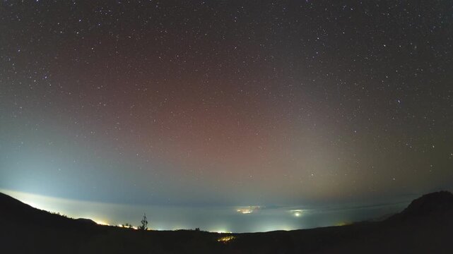 4K timelapse from Tenerife mountains: vibrant zodiacal light glows after sunset, Milky Way arches across dark sky. Ocean horizon below with ships, drifting clouds, volcanic landscape in Teide area