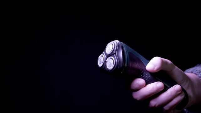 A male hand grips a modern electric rotary shaver against a black backdrop, turning it under dramatic purple light. Side view. Concept of masculine grooming, hygiene technology, and facial care.