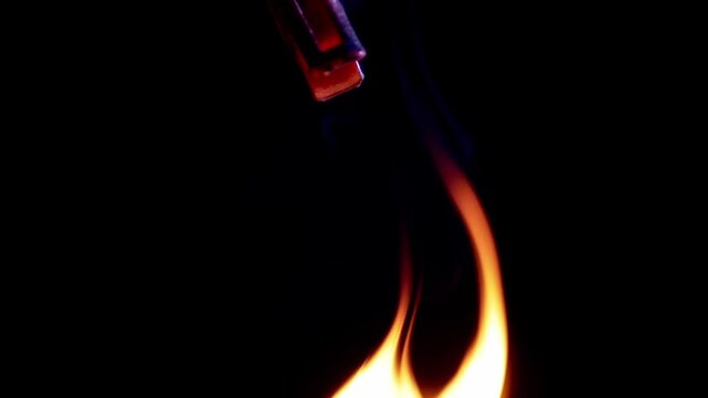 A small object held by a red clip is entirely engulfed in bright orange flames before dropping into complete darkness. Top view. Concept of destruction, burning passion, and transience.